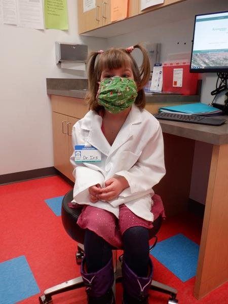 Girl in lab coat and face mask sits in medical office chair; 