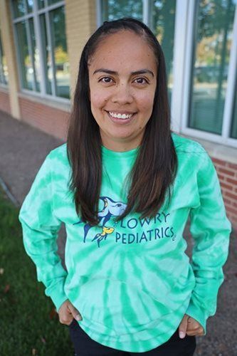 Woman in green tie-dye Lowry Pediatrics shirt smiles outdoors near a building with windows and grass.