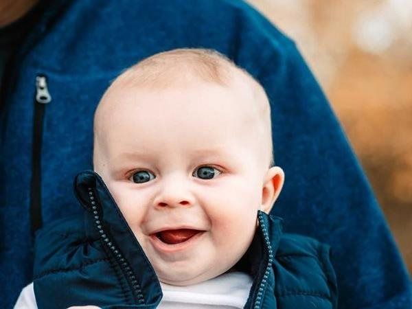 Smiling baby in a blue vest; held by someone wearing a blue jacket, outdoor setting.