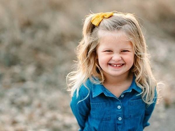 Blonde child smiling broadly, wearing a blue shirt and yellow hair bow, outdoors.