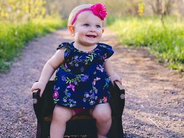 Smiling baby in a blue floral dress and pink headband sitting in a small wooden chair on a path.