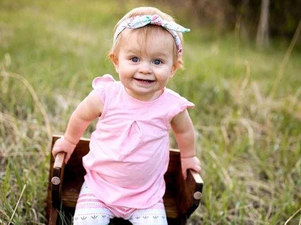 Smiling baby in pink dress and headband sitting in a wooden chair outdoors in tall grass.