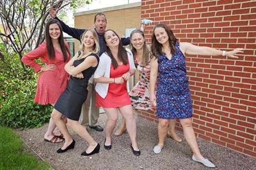 Group of seven people smiling and posing in front of a brick wall, some with arms raised in playful gestures.