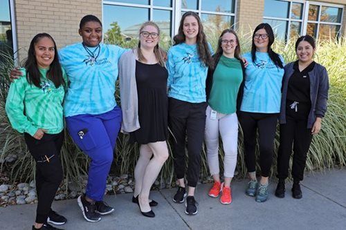 Seven people standing outside, arms around each other. Two wear tie-dye shirts, one a green top, and others in scrubs. Sunny day.