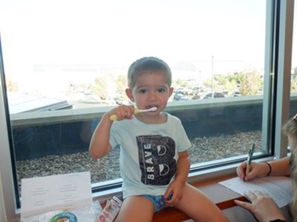 Boy brushing teeth, sitting by a window. He wears a light blue shirt. A woman is seen writing on paper.