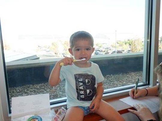 Young child brushing teeth by a window; sunny, neutral setting.