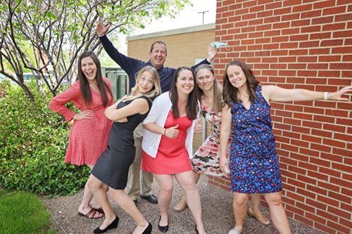 Group of people smiling and posing by a brick wall and bushes.