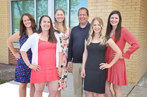 Group of six smiling people posing outdoors by a building; hands on hips.