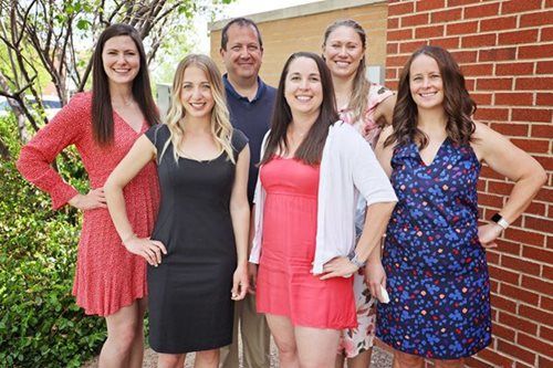 Group of six people smiling, posing by a brick building, wearing casual attire.