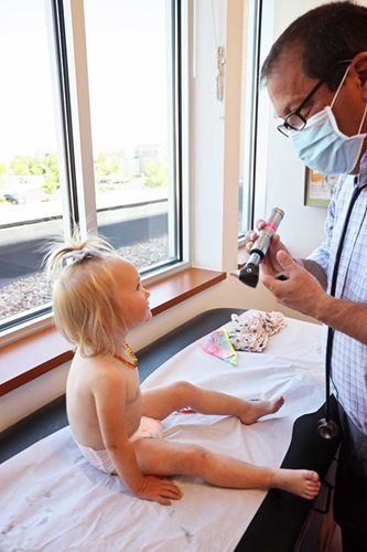 Doctor examining a child's eyes with a light; the child sits on an exam table near a window.