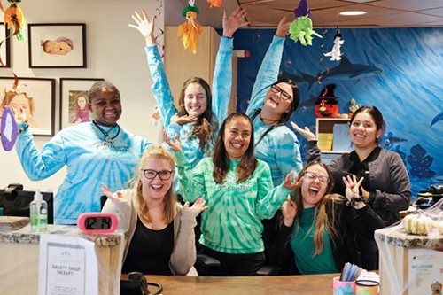 People smiling with arms raised behind a reception desk, wearing tie-dye shirts, decorated room.