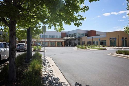 Modern hospital building exterior with parking lot and trees on a sunny day.