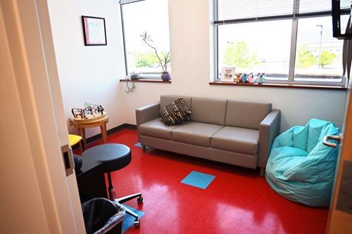 Waiting room with a gray sofa, teal beanbag, red floor, and windows.