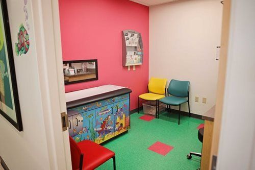 Doctor's office with pink and white walls, colorful floor, child's exam table, and chairs.