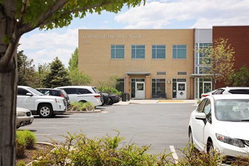 Exterior view of a tan brick building with parking lot. Several cars are parked in front of the building.