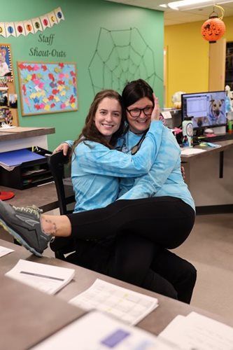 Two women in matching blue shirts embrace at a desk. One sits on the other's lap, both smiling in an office.
