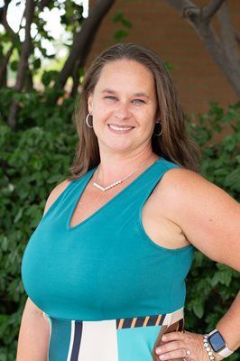 Woman in turquoise top, smiling, posed outdoors near foliage.