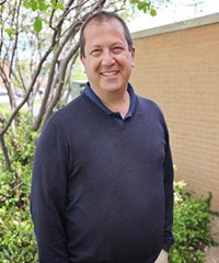 Man smiling, wearing a navy blue sweater, standing outside near a brick wall and bushes.
