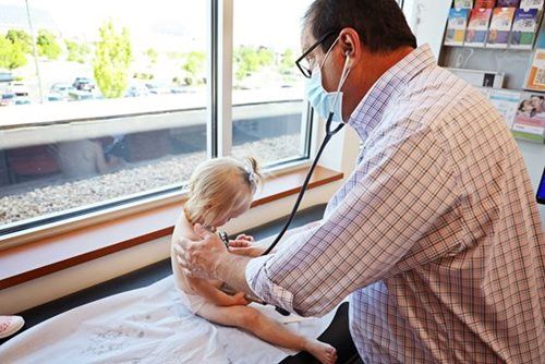 Doctor listening to a child's chest with a stethoscope in a medical office next to a window.