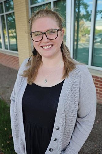 Woman wearing glasses and cardigan smiles outdoors, near brick building with glass windows.