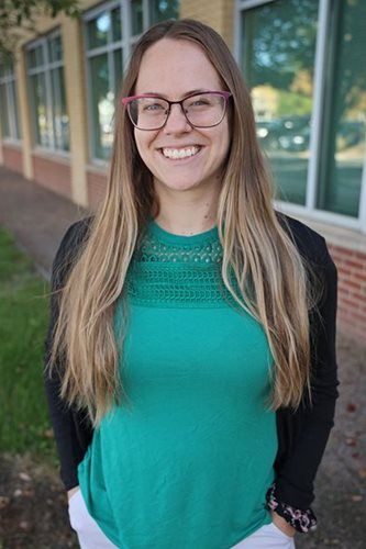 Woman wearing glasses, teal shirt, and cardigan smiles outside a building with windows.
