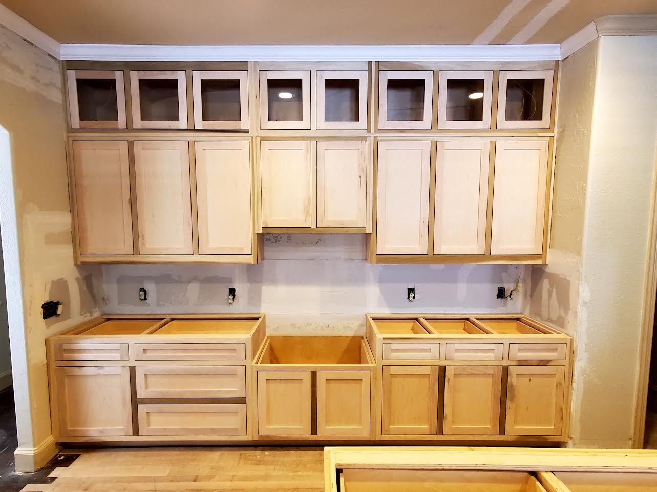 Kitchen cabinets being installed, with upper and lower cabinets in light wood tones against a neutral wall.