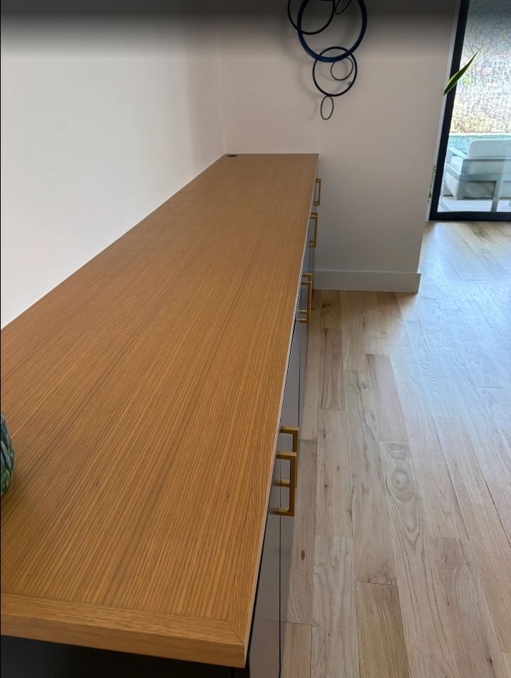 Long, wood-topped cabinet with gold handles, next to light wood flooring and a white wall, under a decorative wall fixture.