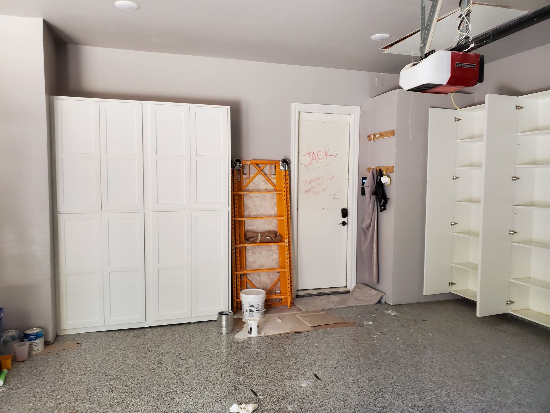 Garage with white cabinets and door. A ladder and paint bucket are in front of the door. The floor is speckled gray.