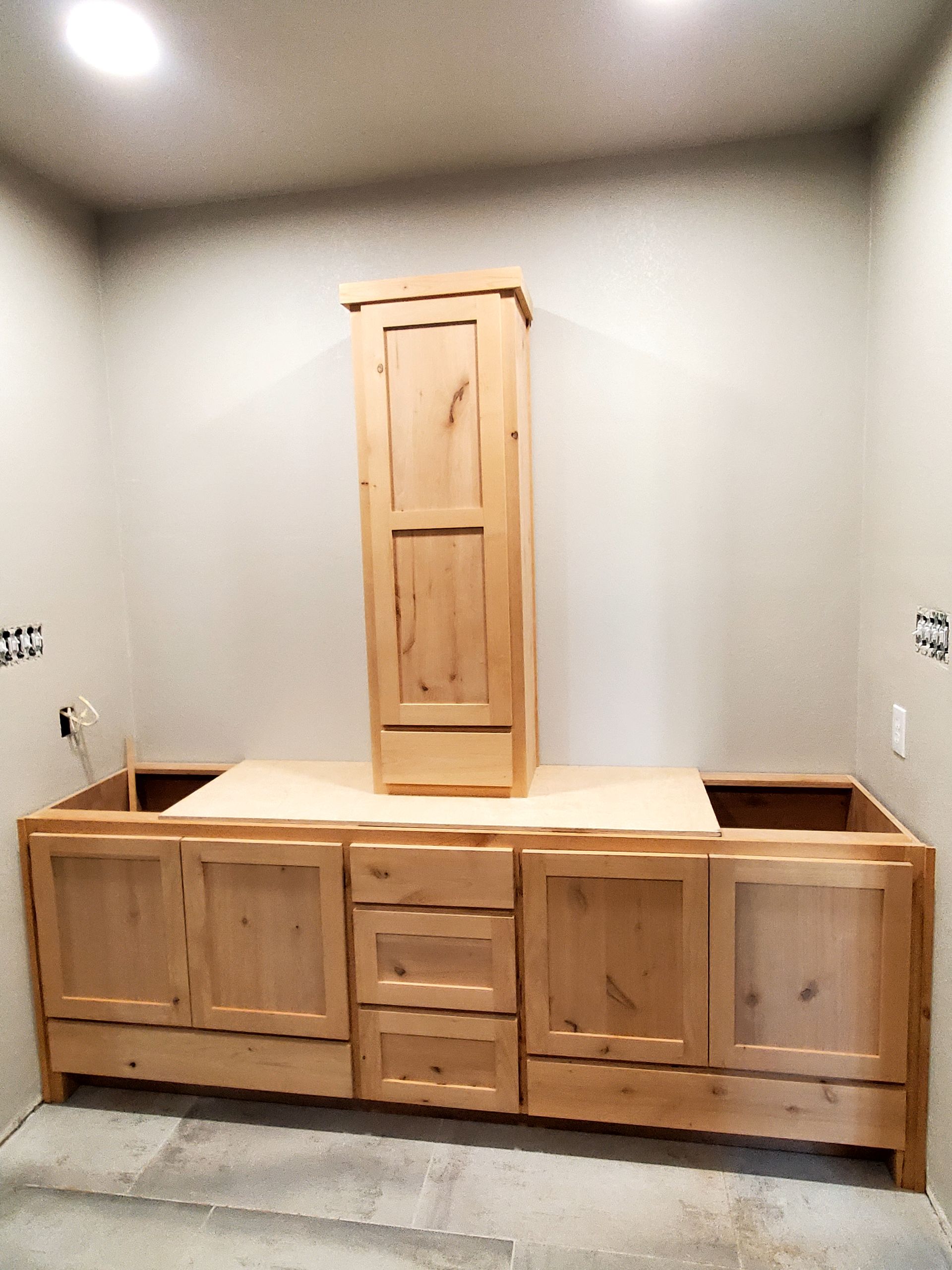 Unfinished wooden bathroom cabinetry being installed. Tall cabinet centered above base cabinets. Gray walls.