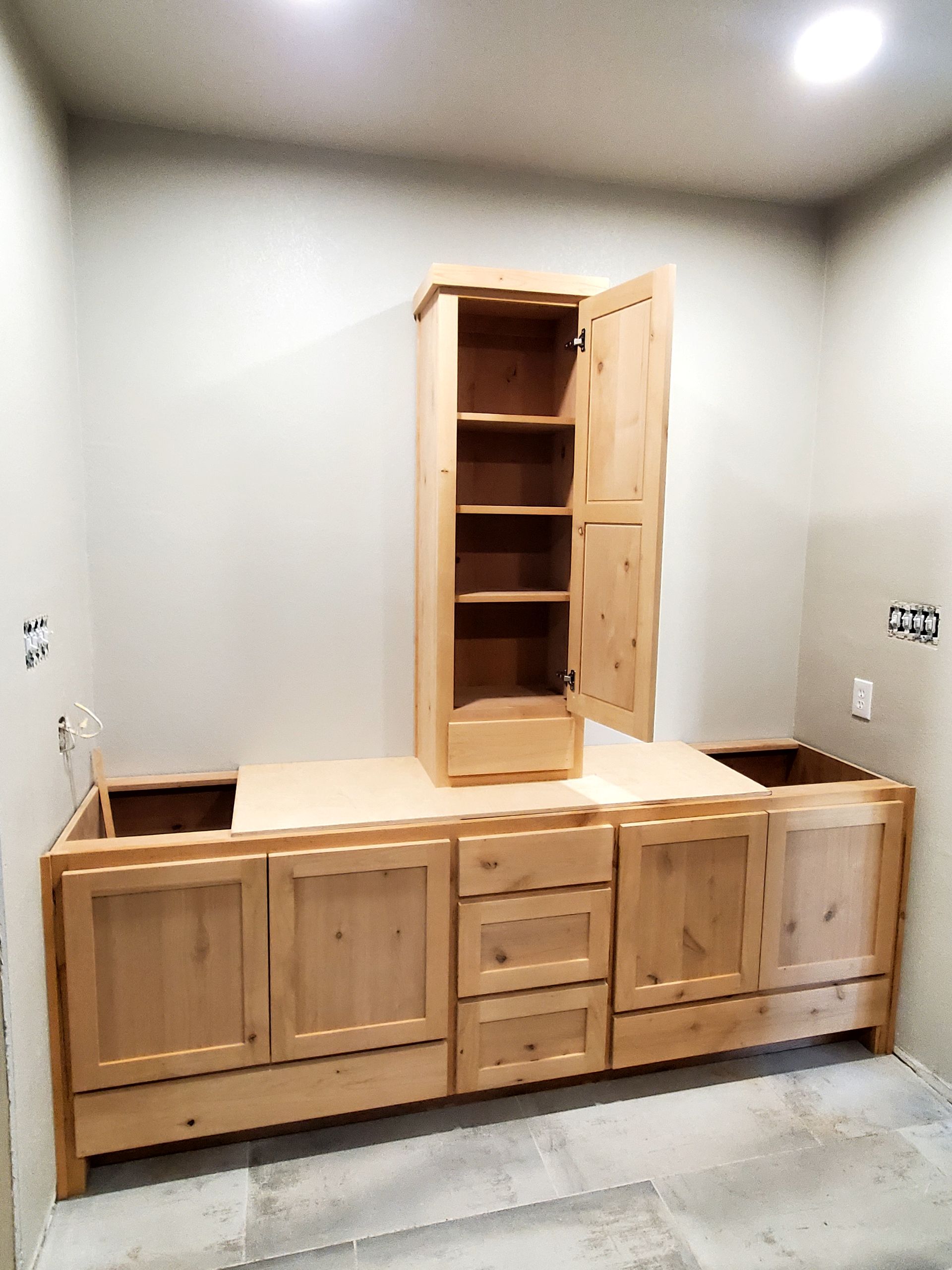Unfinished wooden bathroom cabinets and a tall cabinet against a gray wall. The tall cabinet door is open.
