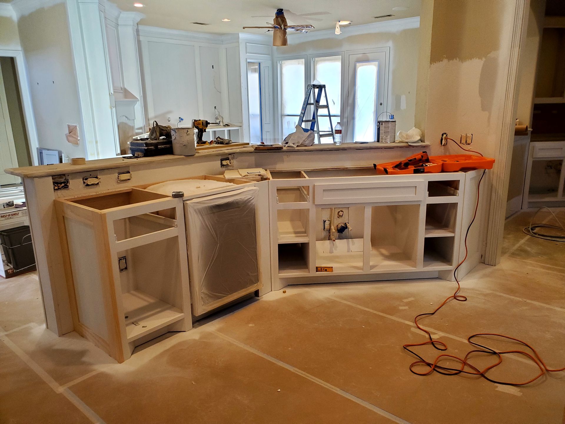 Kitchen under renovation, white cabinets partially assembled, dust on the floor, power tools visible.