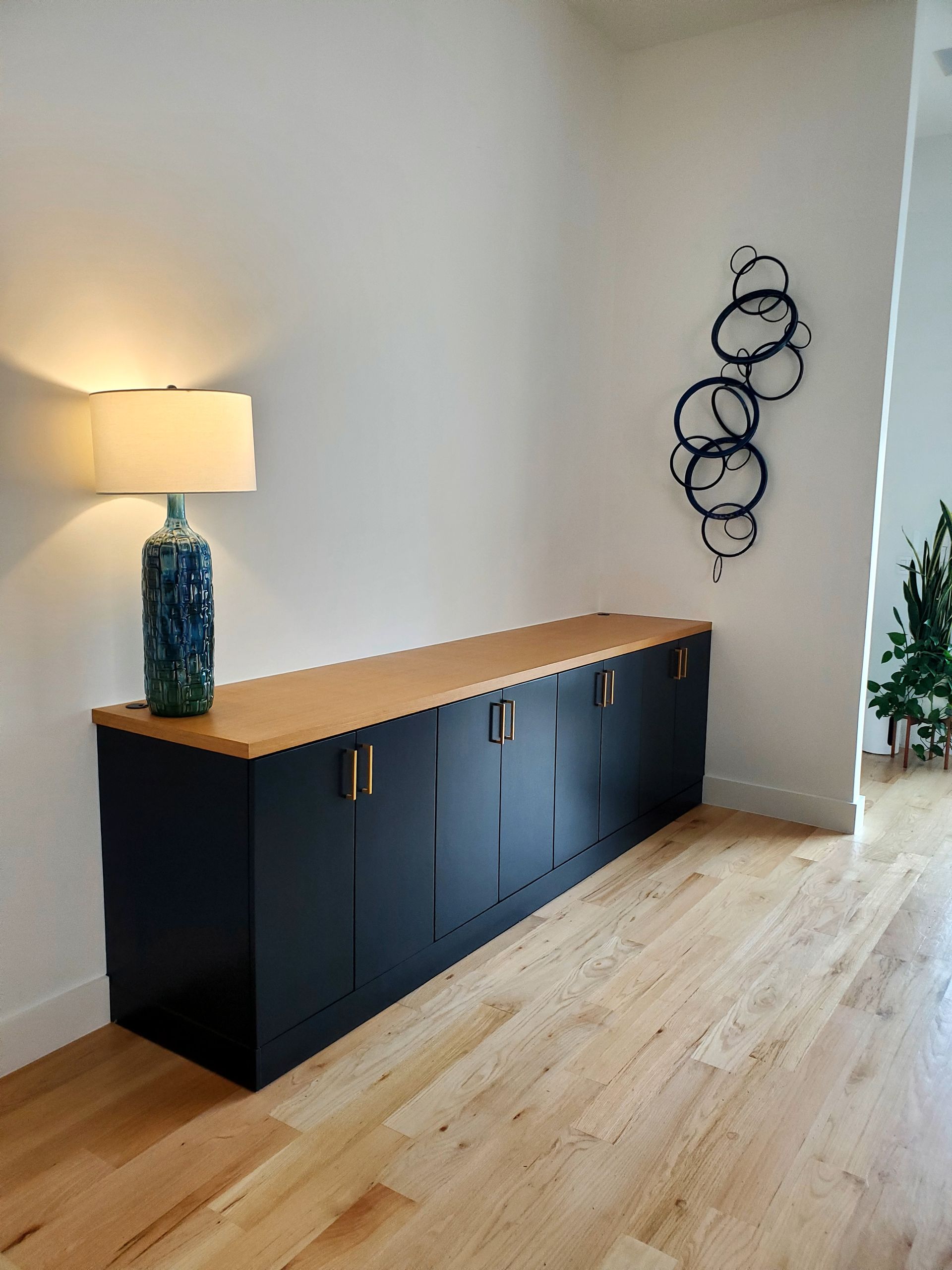 Long dark blue cabinet with wooden top, lamp, and abstract art on white wall.