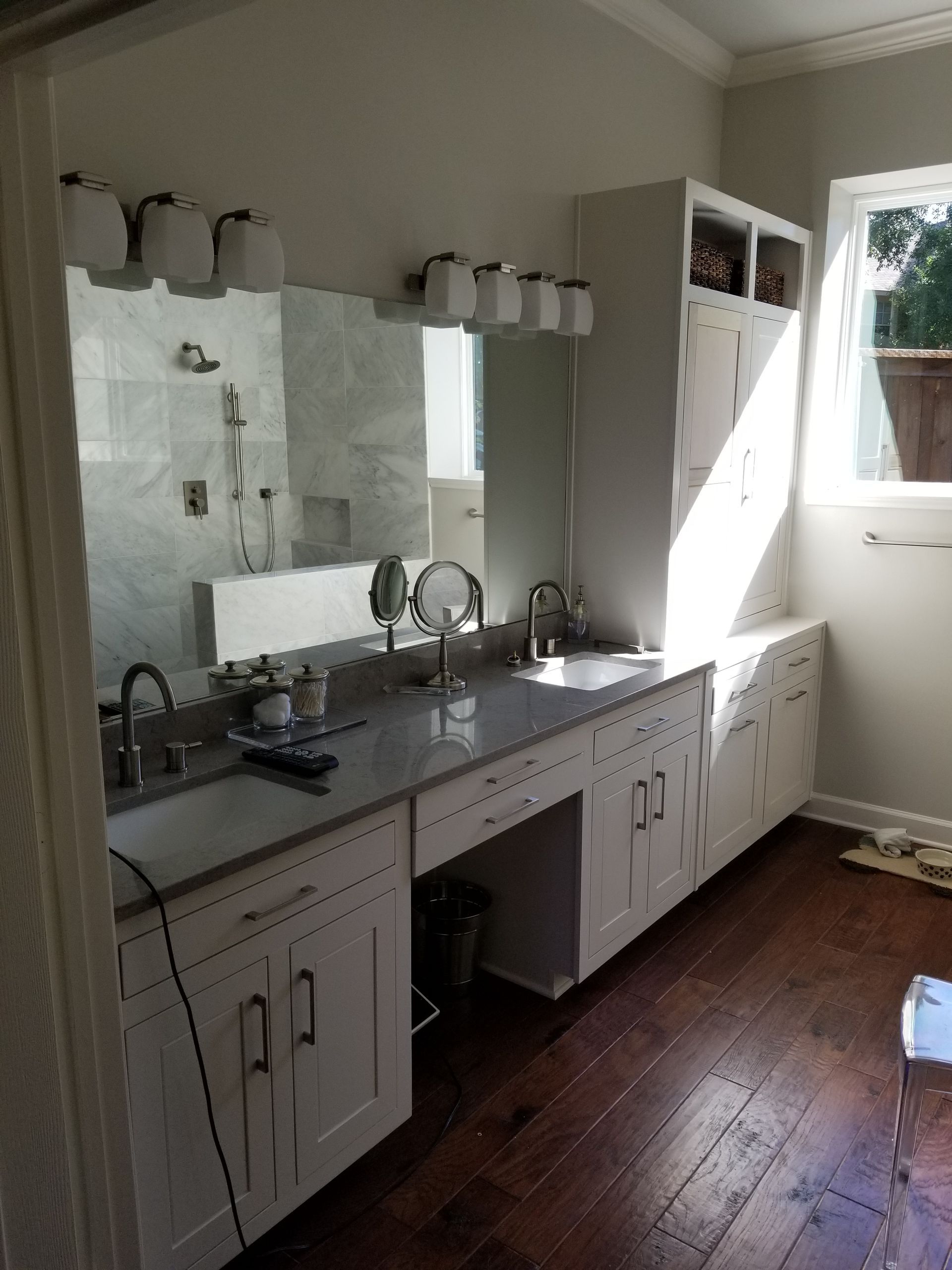 Modern bathroom with white cabinets, gray countertop, and dark wood floors. Double sinks, large mirror, and bright lighting.