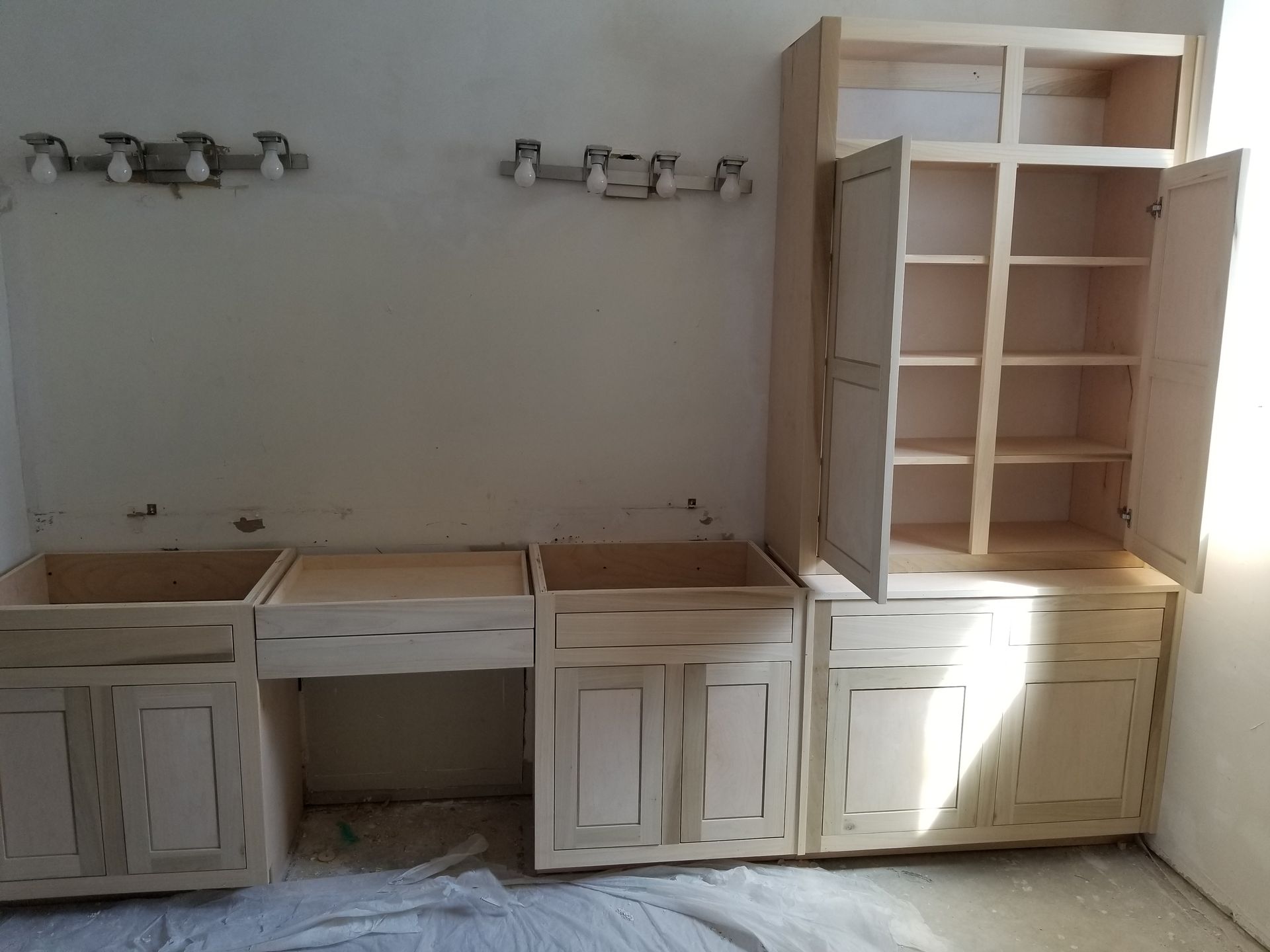 Unfinished wooden kitchen cabinets installed on a wall, with light fixtures above them. The cabinet doors are open.