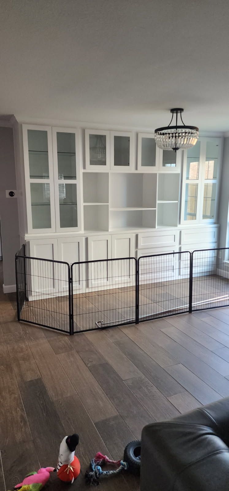A white built-in cabinet with glass doors, a black pet gate in front, and wooden flooring. Toys are scattered on the floor.