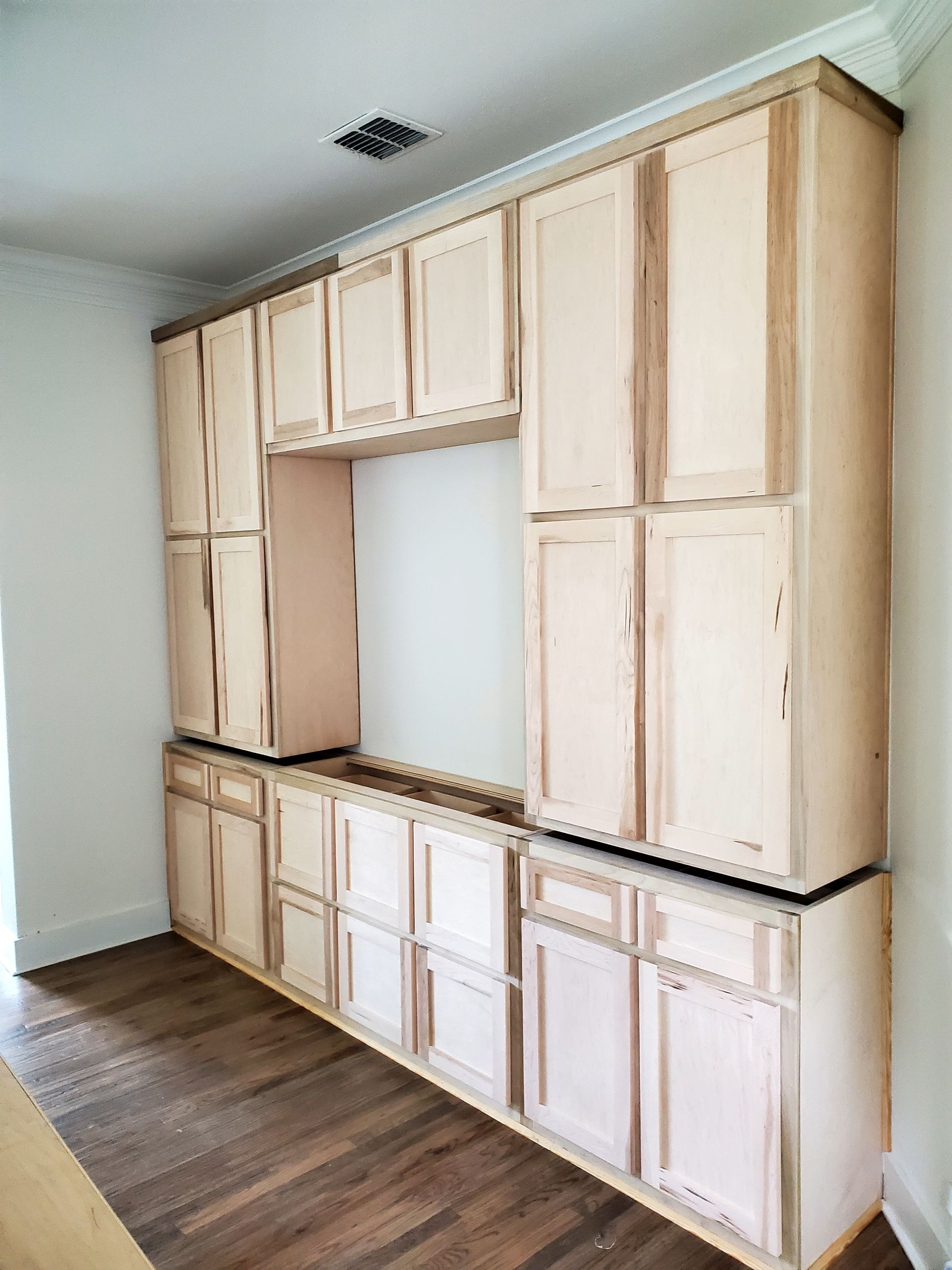 Unfinished wood kitchen cabinets installed on a wall, with upper and lower cabinets surrounding an empty space.