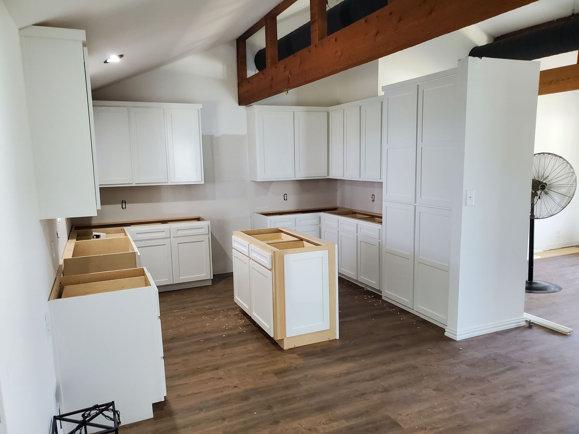 White kitchen cabinets being installed in a room with wood beams and a partially constructed island. Brown flooring.