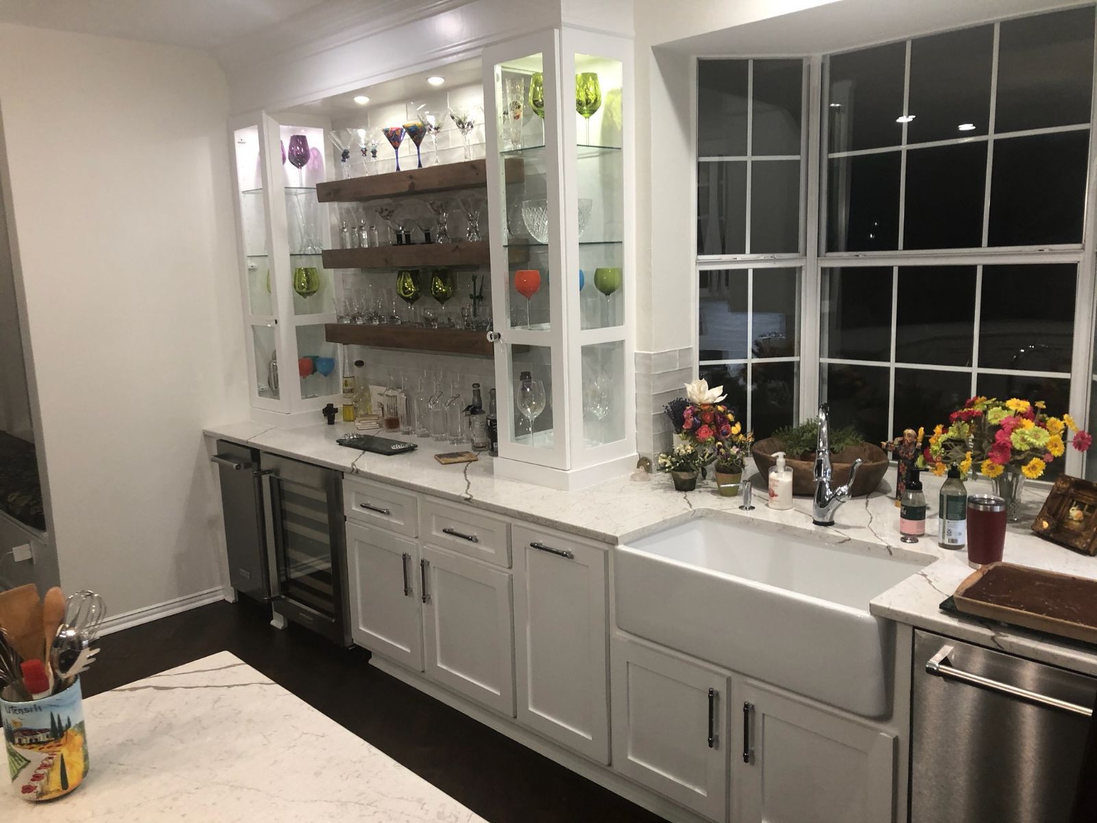 White kitchen with a built-in display cabinet, farmhouse sink, and large window overlooking a dark night.