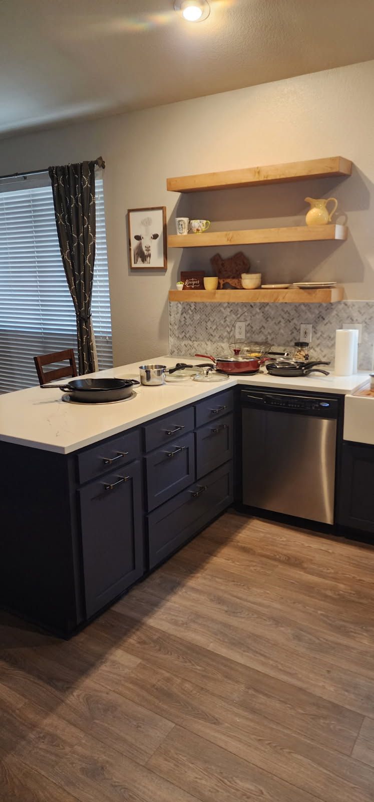 A kitchen with navy blue cabinets, a white countertop, and light wooden shelves. A stainless steel dishwasher is visible.