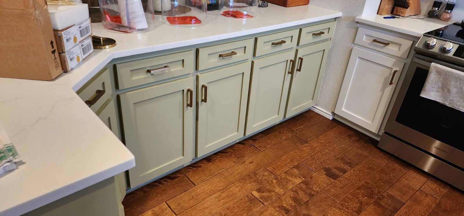 Kitchen cabinets with light green doors and brown hardware, a white countertop, and wooden floors.