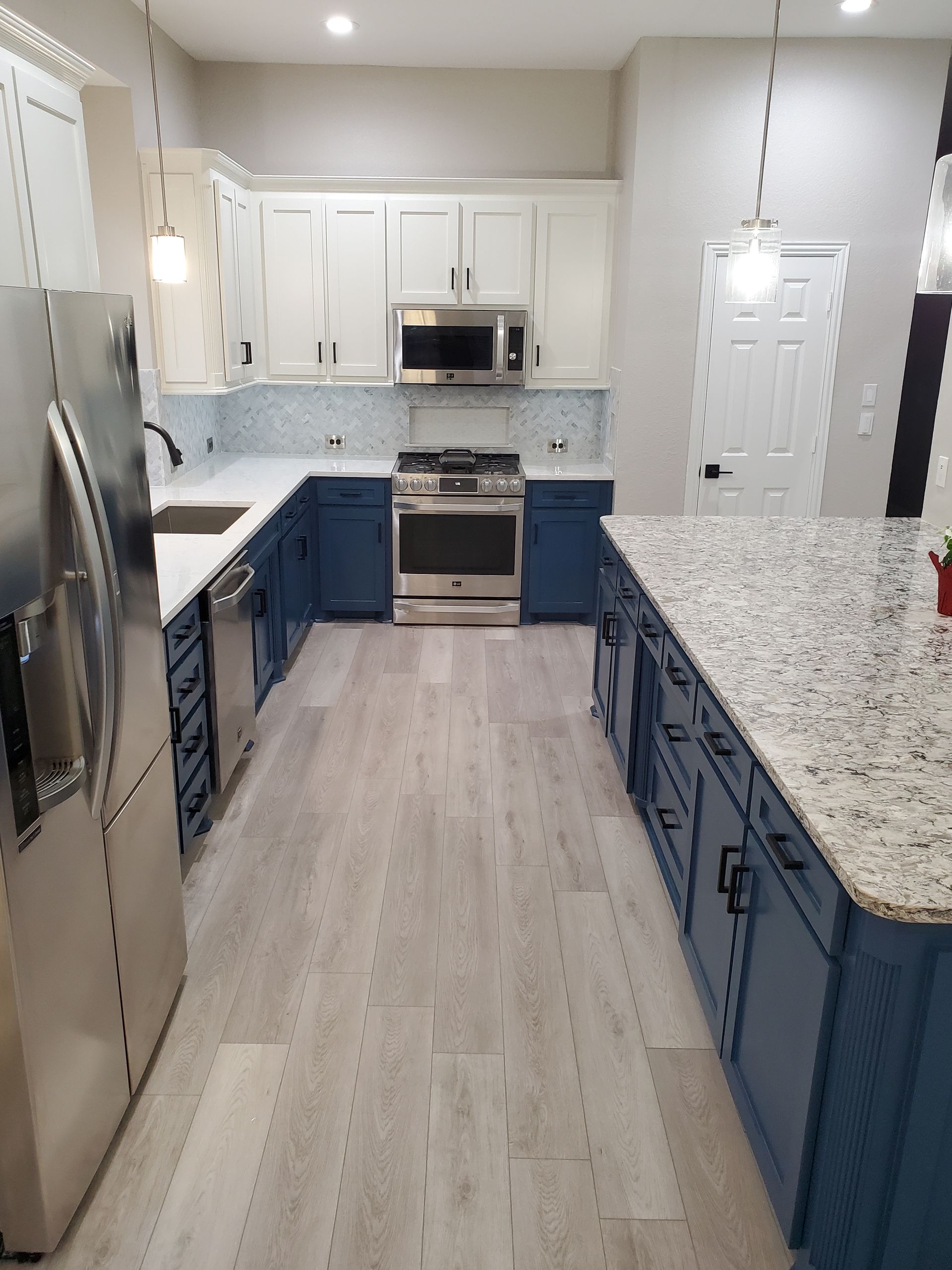 A modern kitchen with white and blue cabinets, stainless steel appliances, and a large island. Light wood-look flooring.