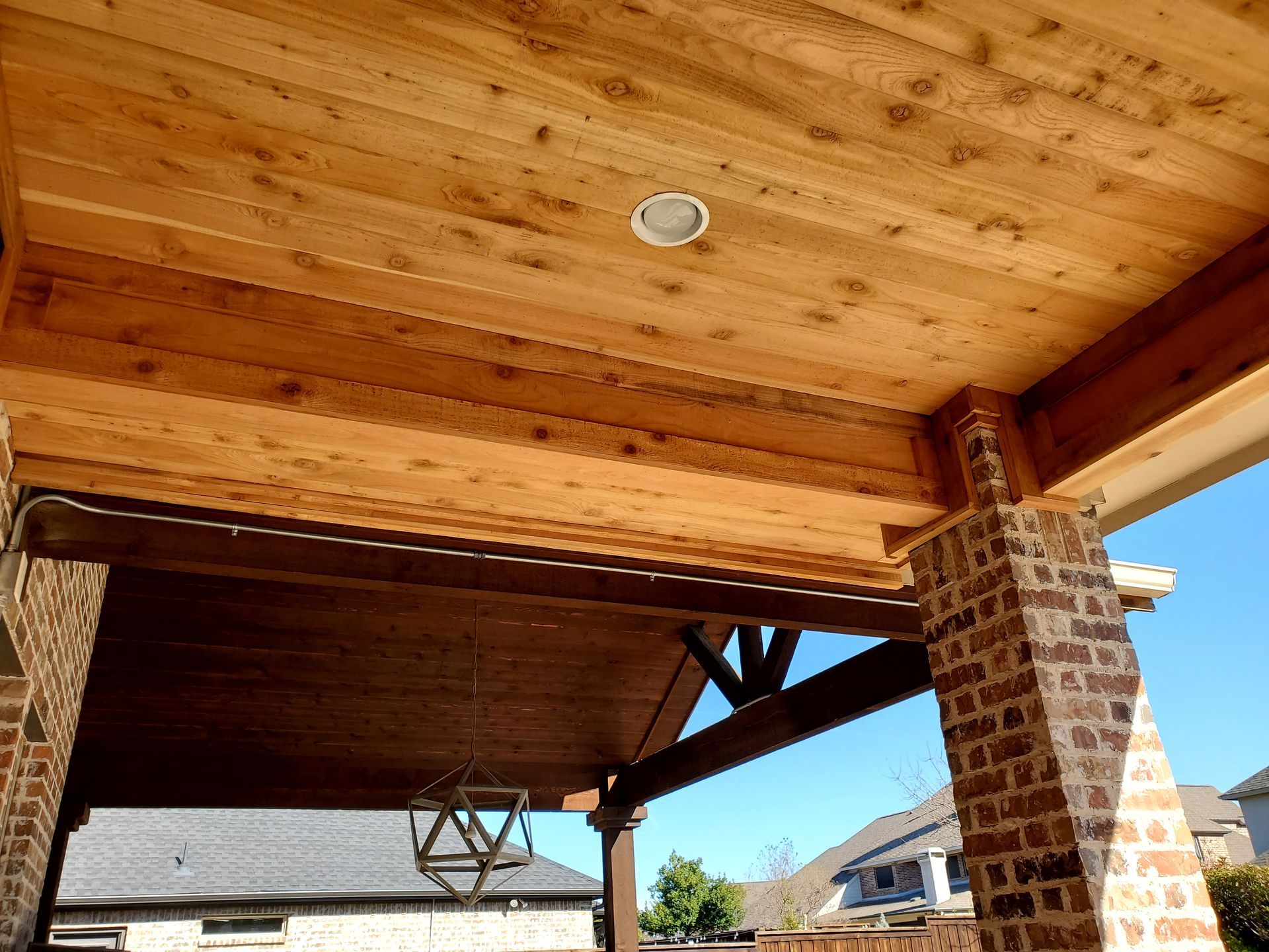 Wooden porch ceiling with a built-in light, supported by brick pillars and wooden beams. Sunny outdoors in the background.