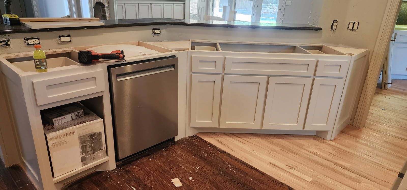 Kitchen cabinets in various stages of installation, featuring a stainless steel dishwasher and white cabinets on a wooden floor.