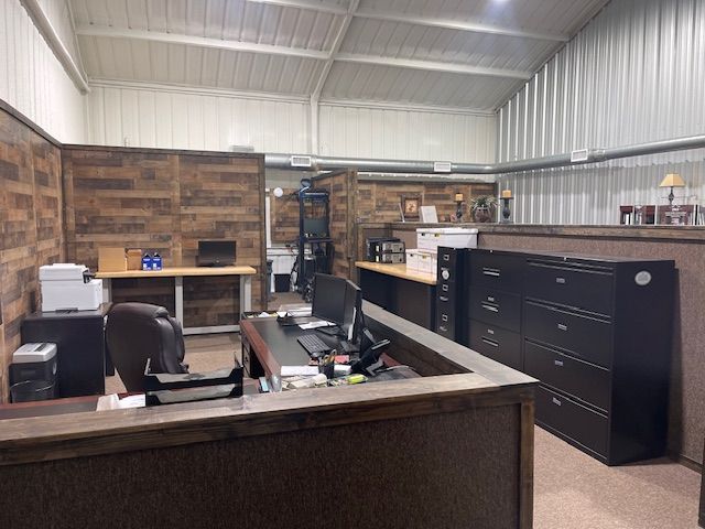 A small office workspace with wood-paneled partitions, black filing cabinets, a desk with a computer, and white metal walls.