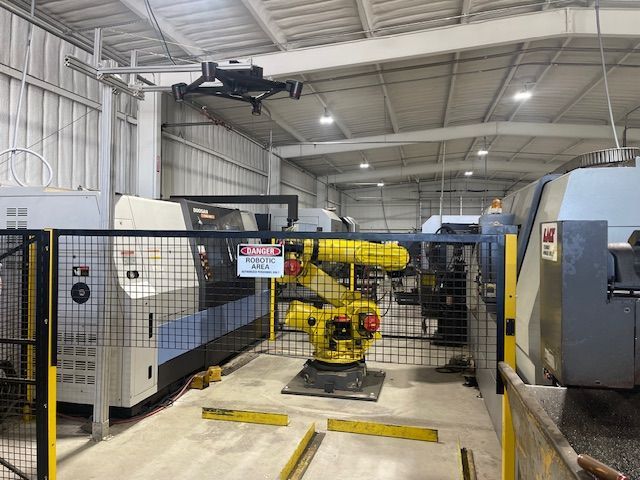 A yellow industrial robotic arm stands behind a safety fence in a manufacturing facility with two machines on either side.