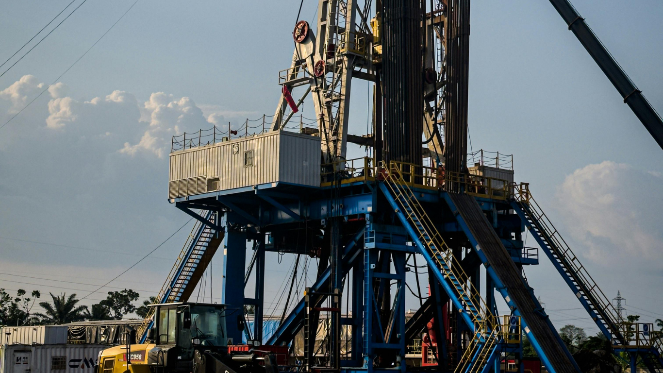An industrial oil drilling rig with a blue support base stands under a partly cloudy sky with a yellow vehicle below.