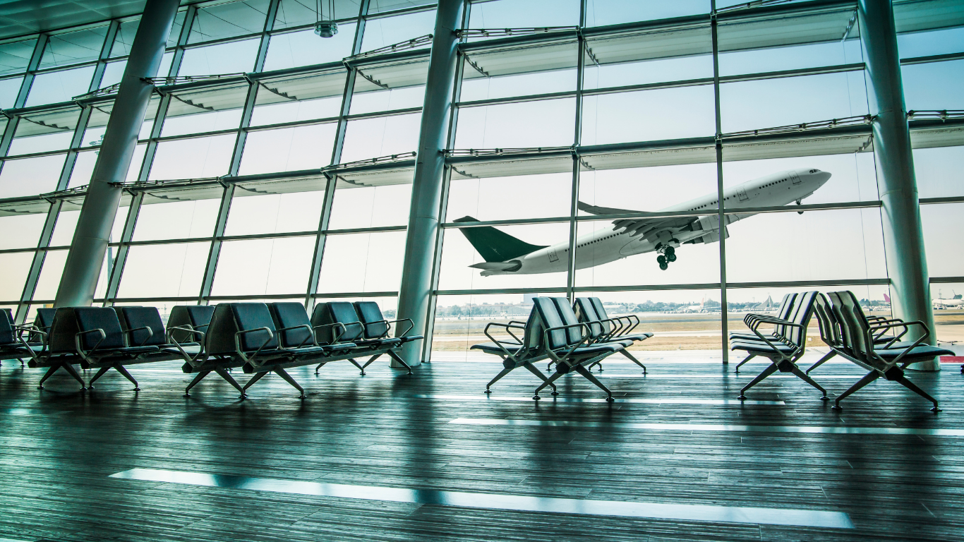 An airplane taking off viewed through the large glass windows of an empty airport terminal with rows of seats.