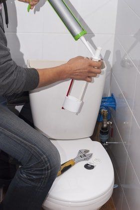 man with caulk fixing bathroom toilet. in small bathroom with white tiles and only a toilet seat. fixing top filtration portion of toilet.