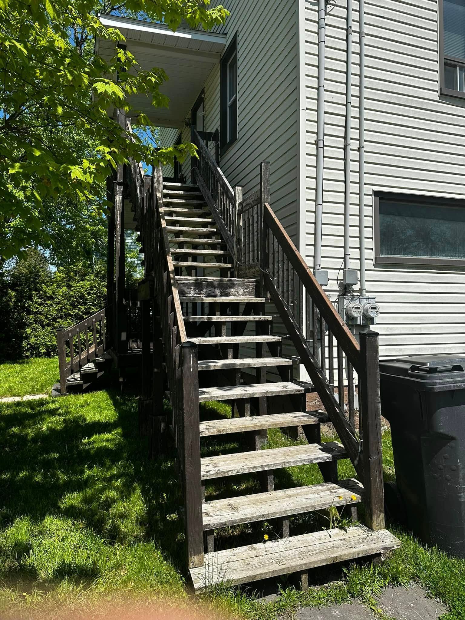 Escalier extérieur en bois avec rambarde, situé à côté d'une maison de couleur claire et menant à une entrée latérale.