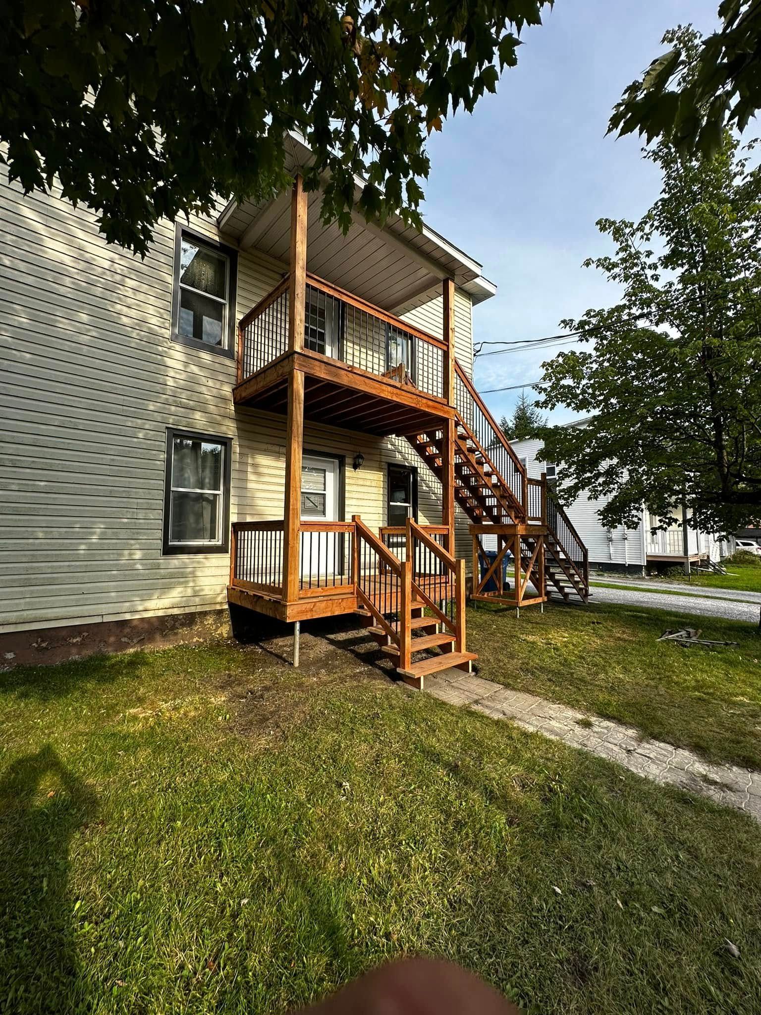 Extérieur d'un immeuble d'appartements avec escalier en bois et porche sous des arbres sur une pelouse.